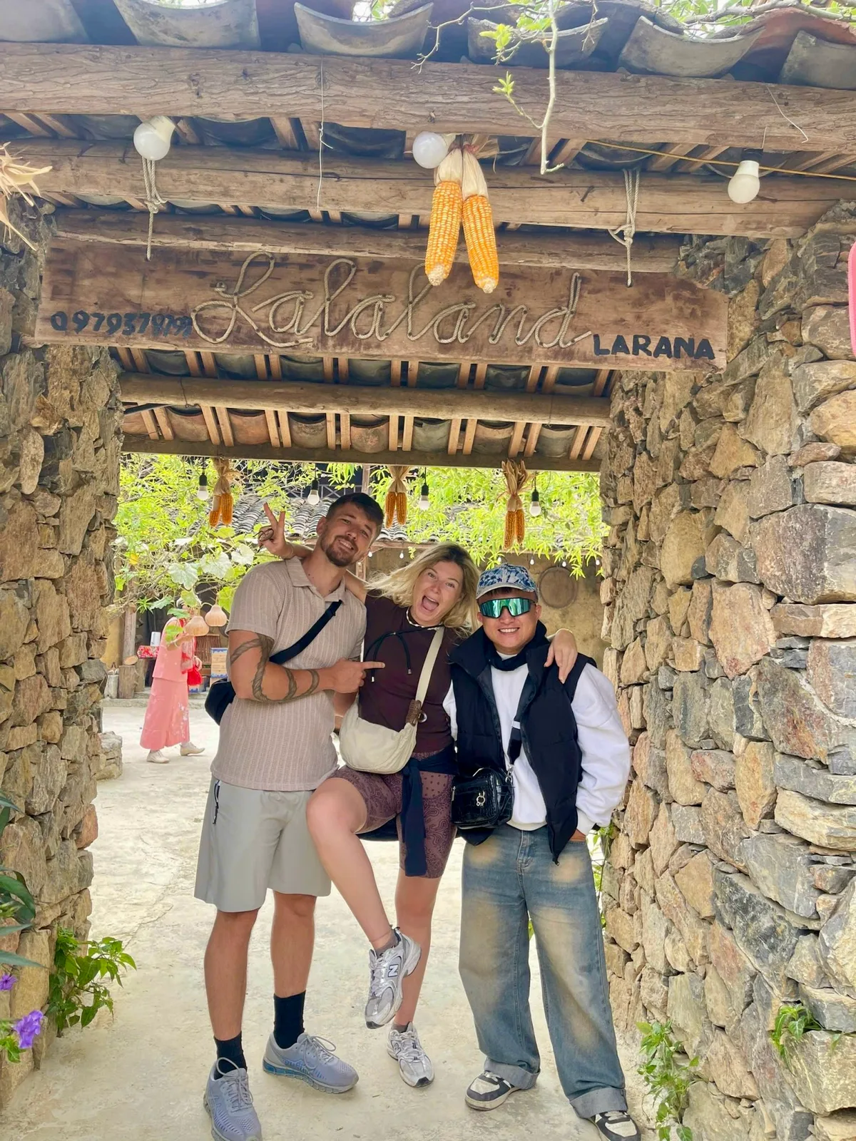 Two guests and one of our team laughing under a stone gateway decorated with dried corn in Ha Giang