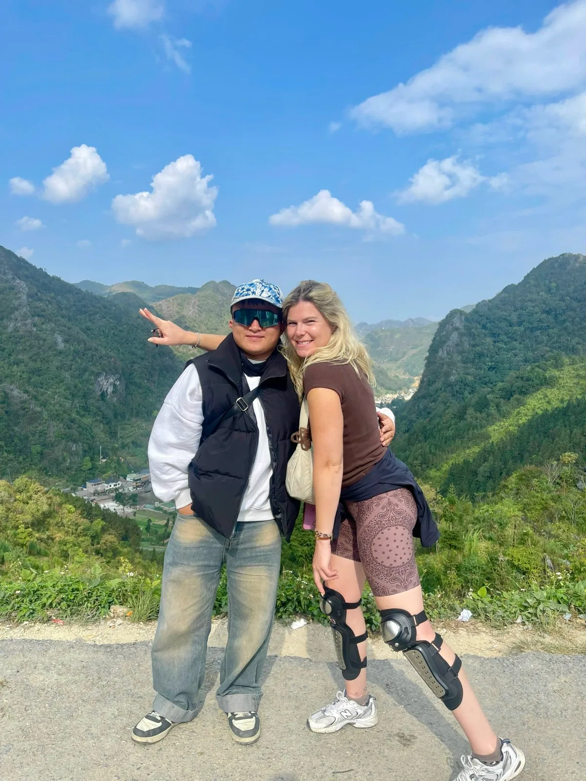 Guest with knee pads and one of our team at a roadside viewpoint over a green valley under blue sky in Ha Giang