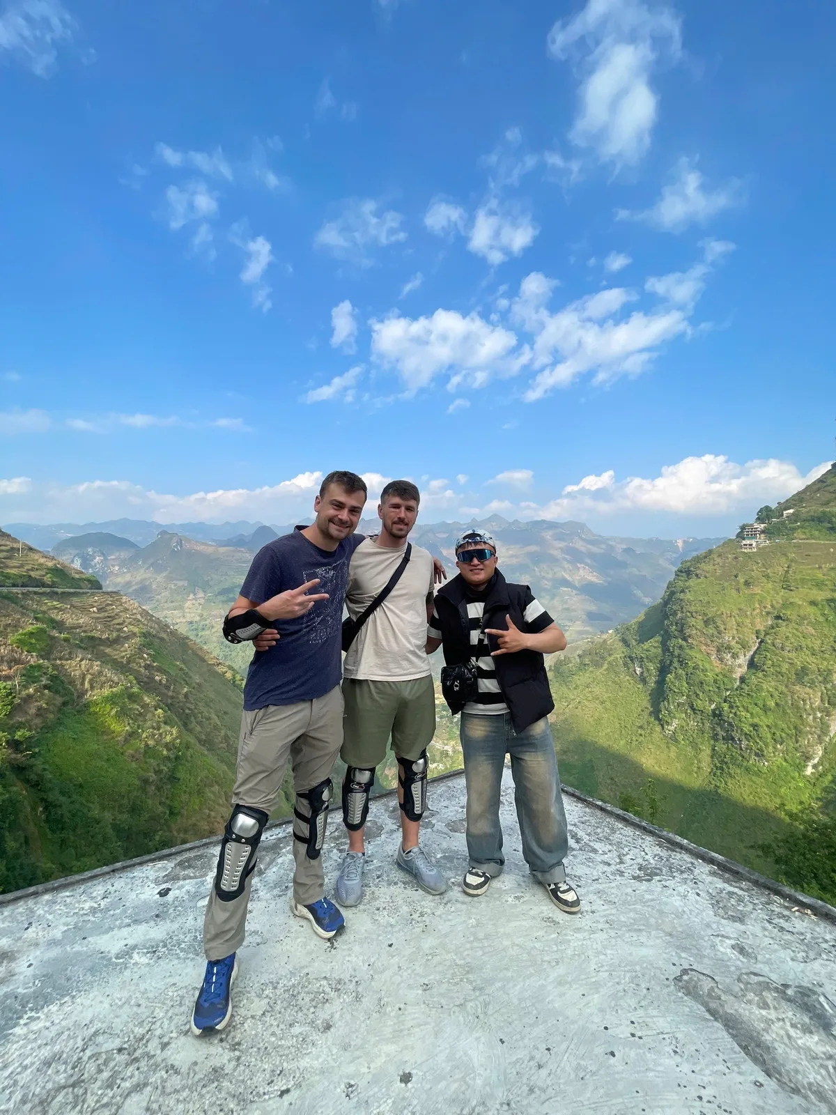 Two guests wearing knee pads with a Freedom team member at a mountain viewpoint under blue sky in Ha Giang
