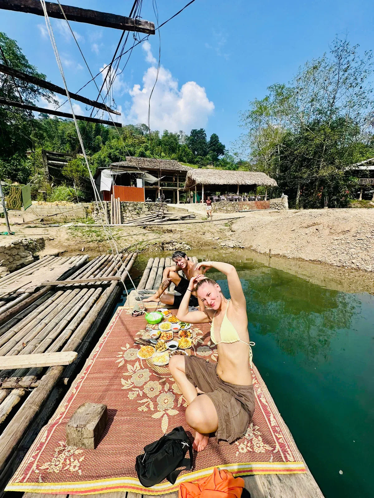 Two guests relaxing beside a tray of lunch dishes on a bamboo raft below thatched houses in Ha Giang