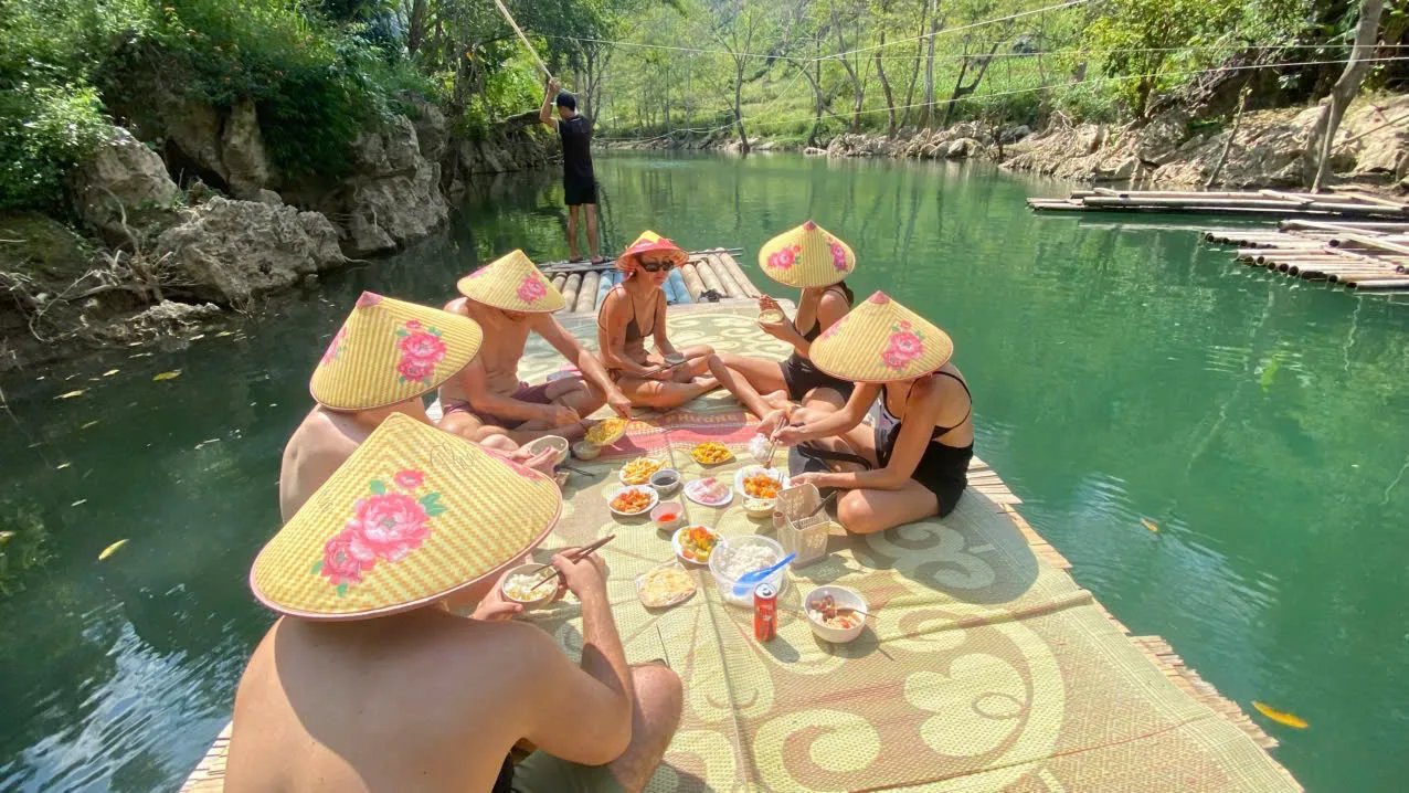 Guests in pink-flowered conical hats eating lunch on a bamboo raft on a green river in Ha Giang
