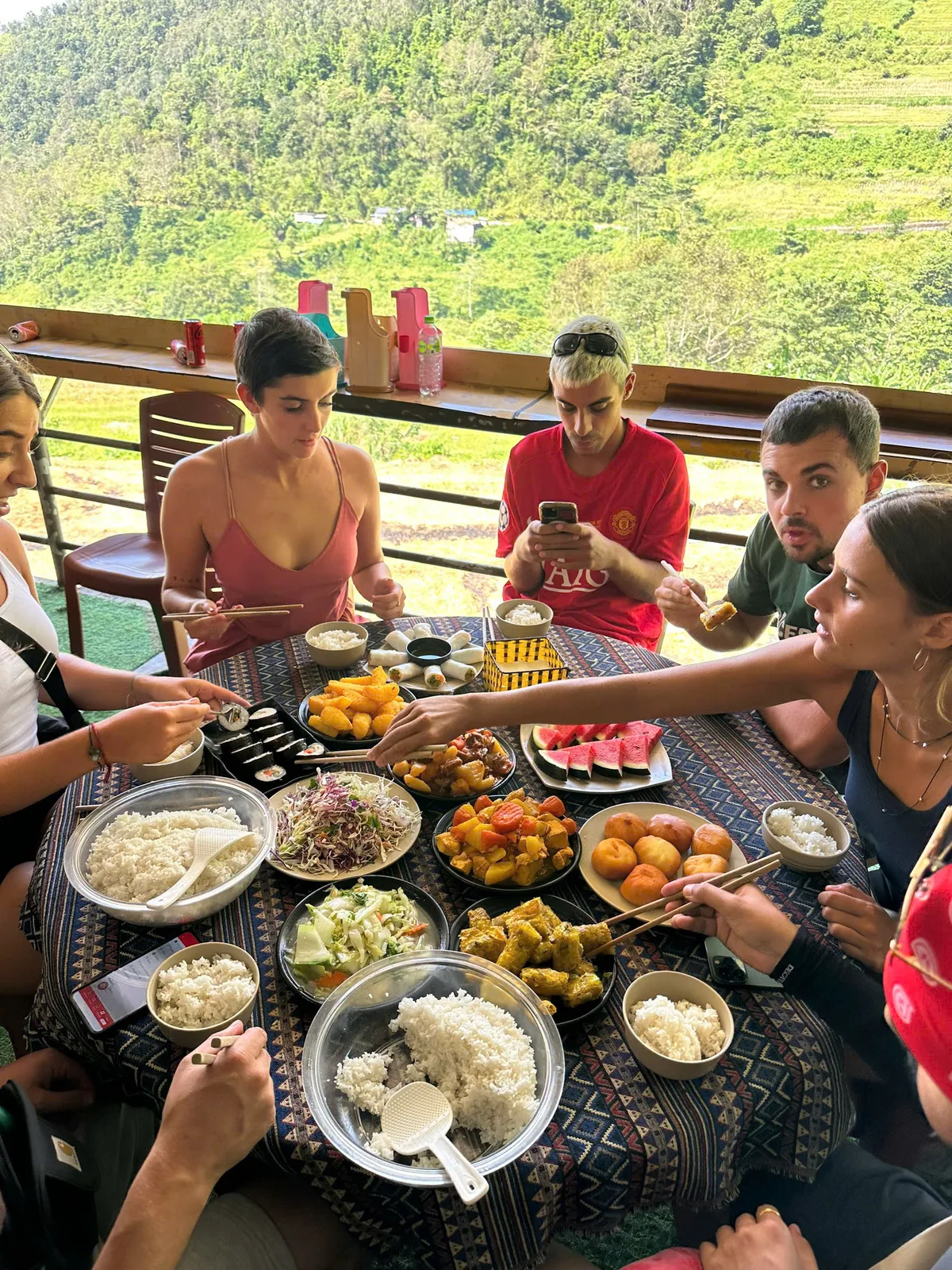 Shared lunch with rice, vegetable dishes, salad, watermelon and spring rolls on a terrace above green hills
